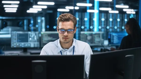 An IT worker in a modern tech workspace monitors multiple computer screens displaying data and system activity.