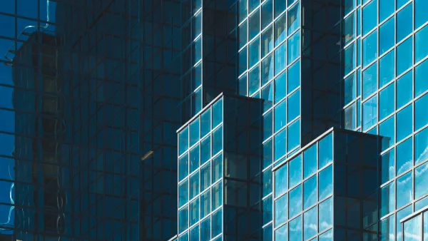 The side of a multi-tiered high rise building made of glass and steel with the reflection of the sky and other buildings in the glass