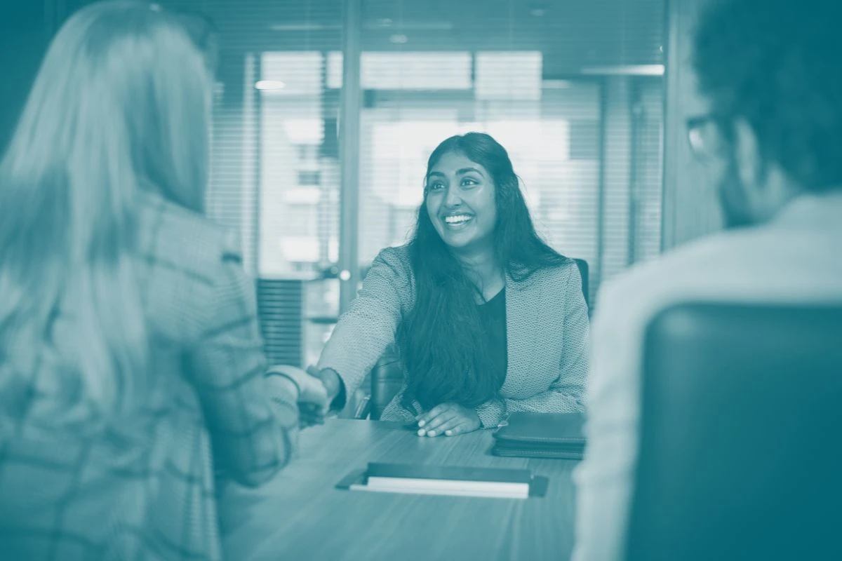 A smiling woman facing the camera and shaking hands with one of two interviewers who are facing away from the camera